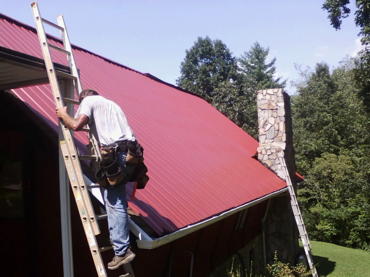 Roofer on a ladder beside a red metal roof and stone chimney — Soffit & Fascia Repair by Roofing Brothers in Carrolltown